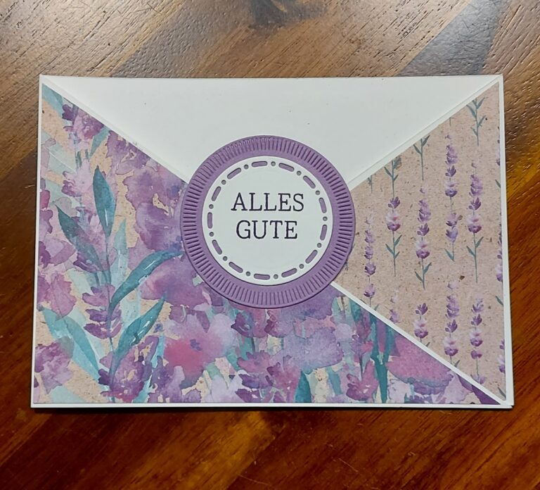 Ein dekorativer Umschlag mit lilafarbenen Blumenmustern und einem kreisförmigen Etikett mit der Aufschrift "ALLES GUTE" liegt auf einer hölzernen Unterlage, perfekt für Ihr Projekt Mach was draus 25. August.