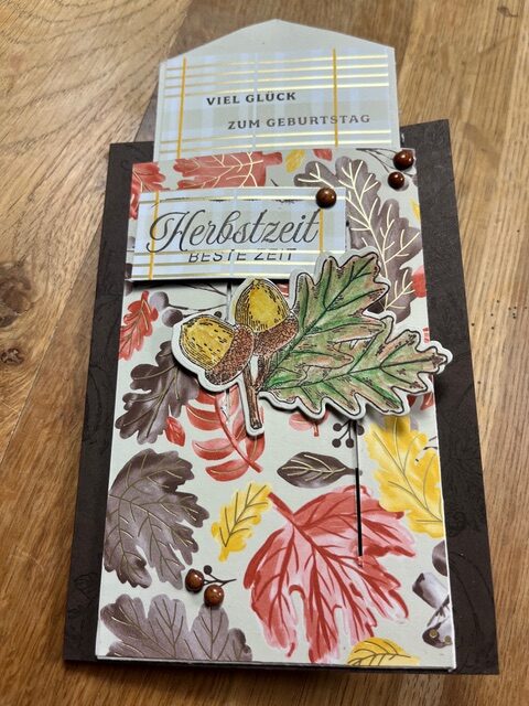 Handgefertigte Herbstkarte mit Eichel- und Blattillustration, deutschem Text "Mach was draus 25. September" und einem gemusterten Hintergrund aus buntem Herbstlaub. Eine zweite Karte ist teilweise in einer Tasche sichtbar.
