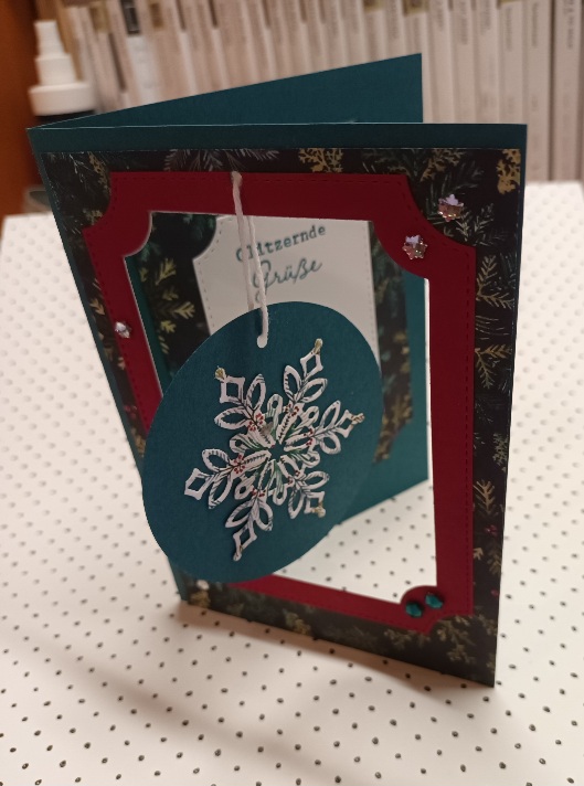 Eine handgefertigte Weihnachtskarte von Mach wurde am 25. Oktober draus, mit einem roten Rand, einem hängenden Schneeflocken-Ornament und einem floral gemusterten Hintergrund, der auf einer weißen gepunkteten Fläche steht.