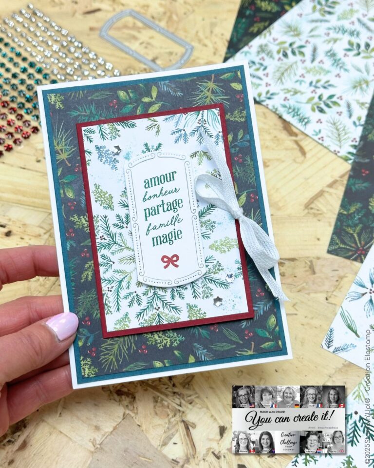 Eine Hand hält eine Feiertagsgrußkarte, die mit grünen Laubmustern, einem weißen Band und französischen Worten verziert ist; "Mach was draus Oktober 25" ist als Teil des Designs inmitten von Bastelmaterial im Hintergrund zu sehen.