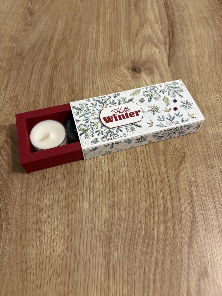 Eine rechteckige Schachtel mit dem Motiv "Hello Winter" aus der Kollektion Mach was draus Oktober 25 liegt teilweise geöffnet auf einer Holzfläche und gibt den Blick auf ein Teelicht frei.