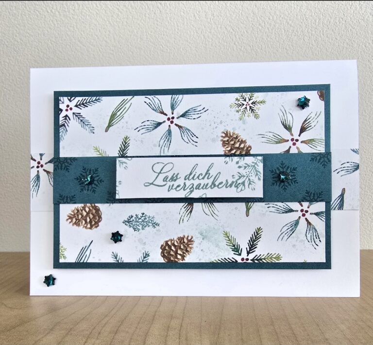 Eine handgefertigte Grußkarte von Mach was draus 25. Oktober zeigt Tannenzapfen- und Tannennadelmuster, blaugrüne Akzente und den deutschen Text "Lass dich verzaubern", der wunderschön auf einer hölzernen Oberfläche dargestellt ist.