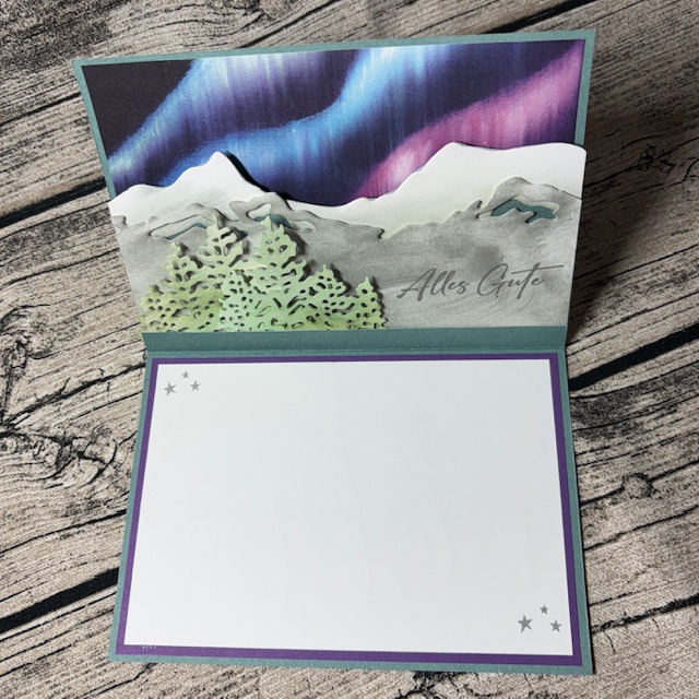 Eine offene Grußkarte von "Mach was draus Oktober 25" mit einer verschneiten Bergszene mit Nordlicht, einem grünen Baum und dem Text "Alles Gute". Der Innenraum ist leer, in lila umrandet mit kleinen Sternen Akzente.
