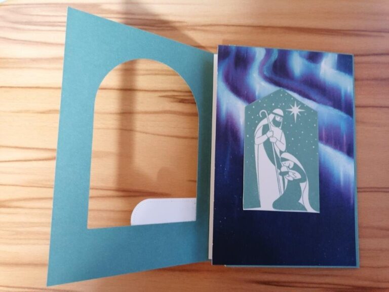 Eine Grußkarte mit einer stilisierten Krippenszene unter einem Stern, mit einem gewölbten Fensterausschnitt auf der linken Seite, einem leuchtenden Hintergrund und der dezent eingearbeiteten Aufschrift "Mach was draus Oktober 25".