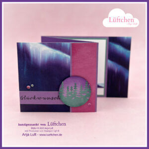 Eine handgefertigte Grußkarte von "Mach was draus Oktober 25" mit einem lila und blauen Aurora-Motiv, dem deutschen Text "Glückwunsch" und einer kreisförmigen Waldsilhouette als Verzierung.