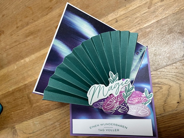Eine handgefertigte Karte von Mach was draus 25. Oktober zeigt einen grünen Papierfächer, den Text "Magic", Blumenmuster und eine deutsche Botschaft, die wunderschön auf einer Holzoberfläche arrangiert sind.