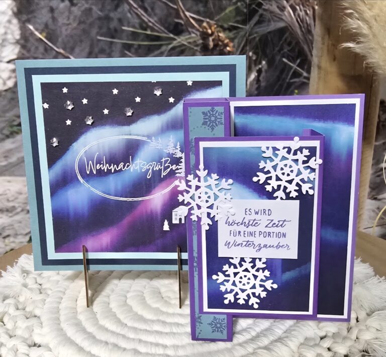 Zwei handgefertigte Weihnachtskarten mit winterlichen Motiven, die für Mach was draus 25. Oktober erstellt wurden, zeigen blaue und violette Aurora-Motive, weiße Schneeflocken und deutsche Texte, die aufrecht auf einer strukturierten Oberfläche stehen.