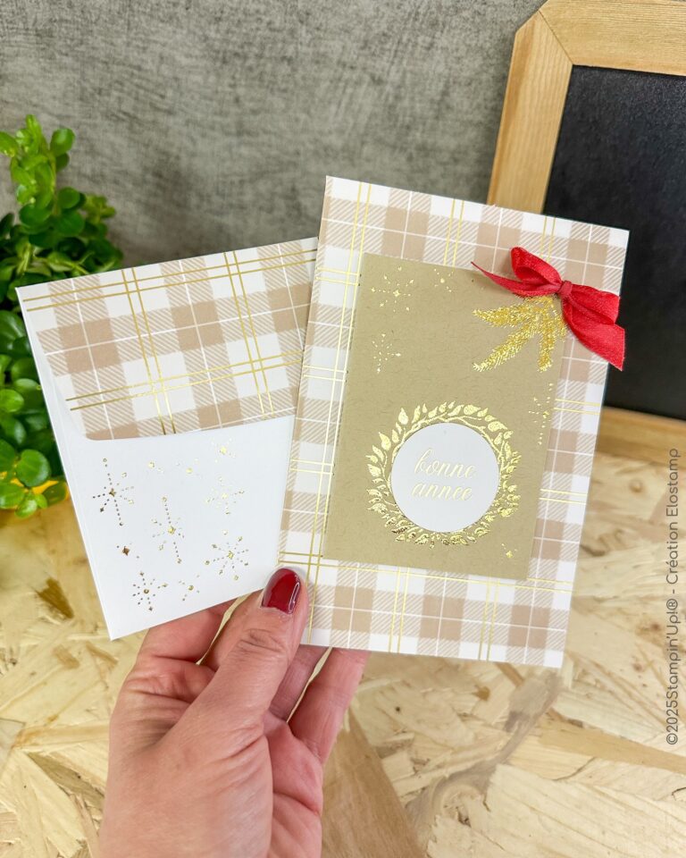 Eine Hand hält eine karierte Grußkarte mit dem Text "bonne année", einer roten Schleife und "Mach was draus Dezember 25" neben einem passenden Umschlag mit goldenen Sternen.