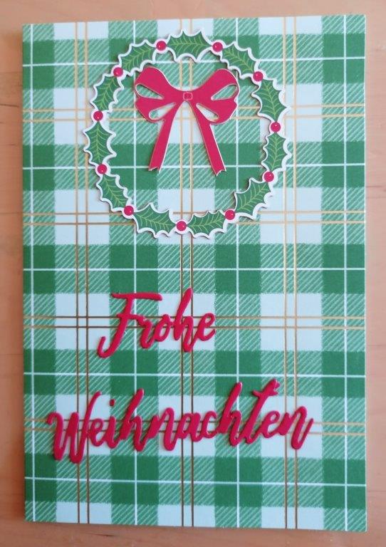 Grün-weiß karierte Karte mit goldenem Gittermuster, Teil von "Mach was draus Dezember 25", mit einem Stechpalmenkranz mit roter Schleife und den Worten "Frohe Weihnachten" in roter Schrift.