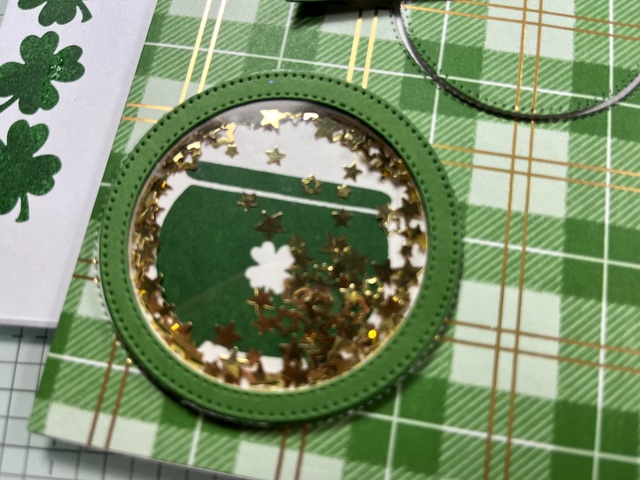 Eine kreisförmige grüne Karte mit einem Topf aus Gold, gefüllt mit goldenem Konfetti in Sternform, auf einem grün karierten Hintergrund - perfekt für die Feierlichkeiten von Mach was draus am 25. Dezember.