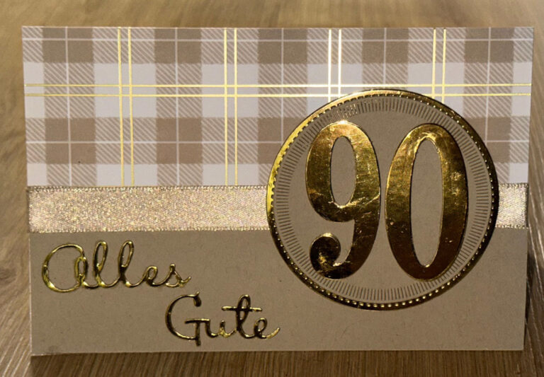 Eine Grußkarte von Mach was draus 25. Dezember zeigt ein kariertes Muster, ein goldenes Band und eine große goldene "90" neben den deutschen Worten "Alles Gute" auf einer Holzoberfläche.