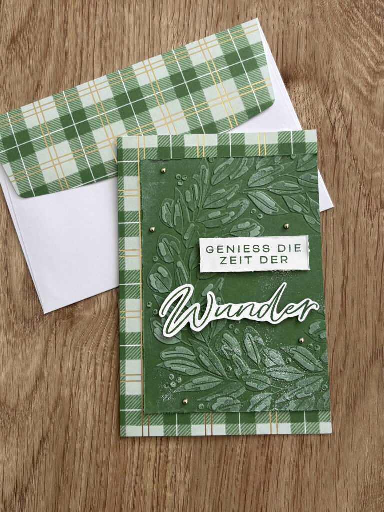 Eine grüne Grußkarte mit Blattmustern und dem deutschen Text "GENIESS DIE ZEIT DER WUNDER", perfekt für Mach was draus 25. Dezember, in einem passenden grün karierten Umschlag auf einer Holzfläche.