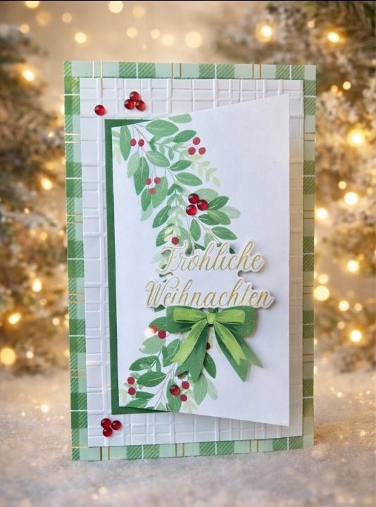 Eine handgefertigte Weihnachtskarte von "Mach was draus Dezember 25" zeigt grünes Laub, rote Beeren, eine grüne Schleife und "Fröhliche Weihnachten" vor einem unscharfen Hintergrund aus Weihnachtsbeleuchtung und einem Baum.