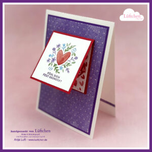 Eine handgefertigte Grußkarte mit einem Herz- und Blumendesign, mit dem deutschen Text "Fühl dich fest gedrückt" auf einer geneigten quadratischen Vorderseite, inspiriert von Mach was draus Januar 2026, mit einem lila Hintergrund und einem Herzmuster innen.