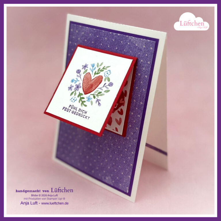 Eine handgefertigte Grußkarte mit einem Herz- und Blumendesign, mit dem deutschen Text "Fühl dich fest gedrückt" auf einer geneigten quadratischen Vorderseite, inspiriert von Mach was draus Januar 2026, mit einem lila Hintergrund und einem Herzmuster innen.