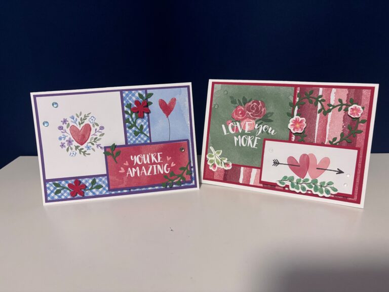 Zwei handgefertigte Grußkarten mit Herz- und Blumendesigns, inspiriert von Mach was draus 26. Januar, eine mit der Aufschrift "You're Amazing" und die andere mit "Love You More", dargestellt auf einer weißen Fläche vor einem dunkelblauen Hintergrund.