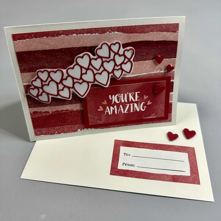 Eine Grußkarte mit roten und rosafarbenen Streifen, Herzverzierungen und der Botschaft "You're Amazing", abgebildet mit einem passenden Umschlag mit der Aufschrift "To" und "From" - perfekt für Mach was draus 26. Januar!.