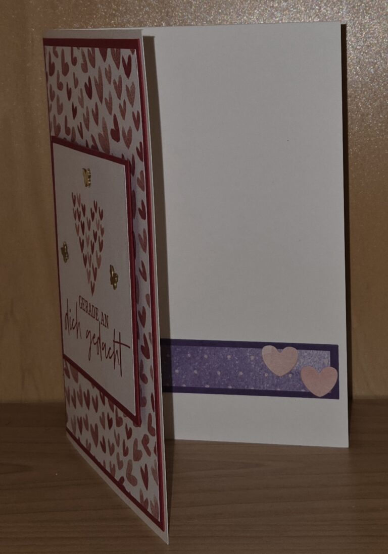 Eine handgefertigte Grußkarte steht offen auf einer Holzfläche. Die Vorderseite zeigt rote Herzen, einen deutschen Text und "Mach was draus 26. Januar", während die Innenseite mit einem lila Streifen und zwei rosa Herzen verziert ist.