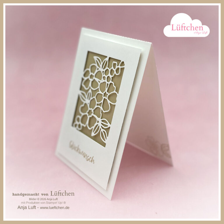 Eine handgefertigte weiße Grußkarte steht aufrecht, mit einem lasergeschnittenen Blumenmuster und dem Wort "Glückwunsch" auf der Vorderseite. Exklusiv online erhältlich ab 26. Januar. Der Hintergrund ist neutral mit einem weichen Fokus.