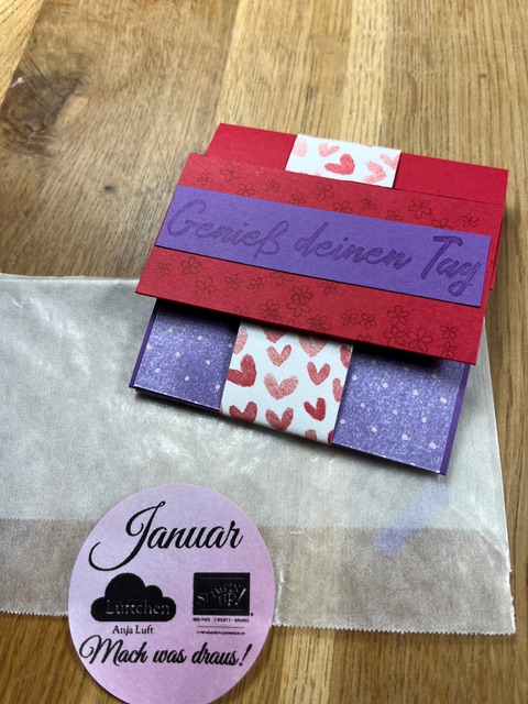 Eine handgefertigte Karte mit dem Text "Genieß deinen Tag" liegt auf einem Holztisch, daneben ein durchsichtiger Umschlag und ein runder Aufkleber mit der Aufschrift "Januar". Perfekt für Mach was draus Januar 26 Projekte.