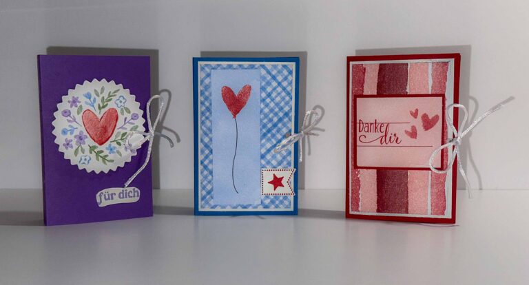 Drei handgefertigte Grußkarten mit Herzmotiven und deutschem Text, inspiriert von Mach was draus 26. Januar, jeweils mit einer weißen Schleife gebunden, stehen aufrecht auf einer weißen Fläche.