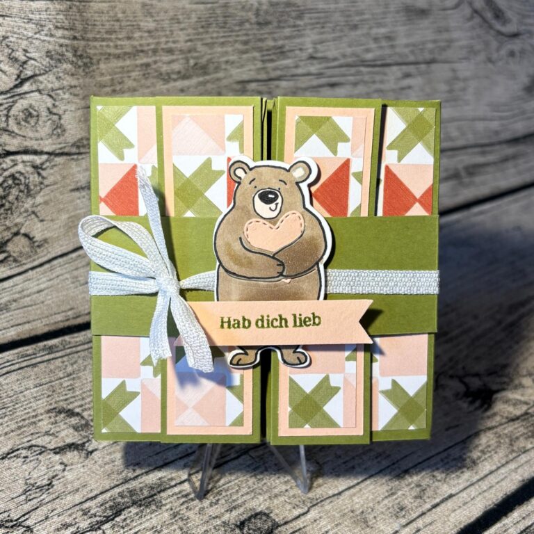Eine handgefertigte Grußkarte, inspiriert von Mach was draus 26. Februar, zeigt einen Bären, der ein Herz hält, geometrisch gemusterte Felder, ein weißes Band und ein "Hab dich lieb"-Banner auf einem rustikalen Holzhintergrund.