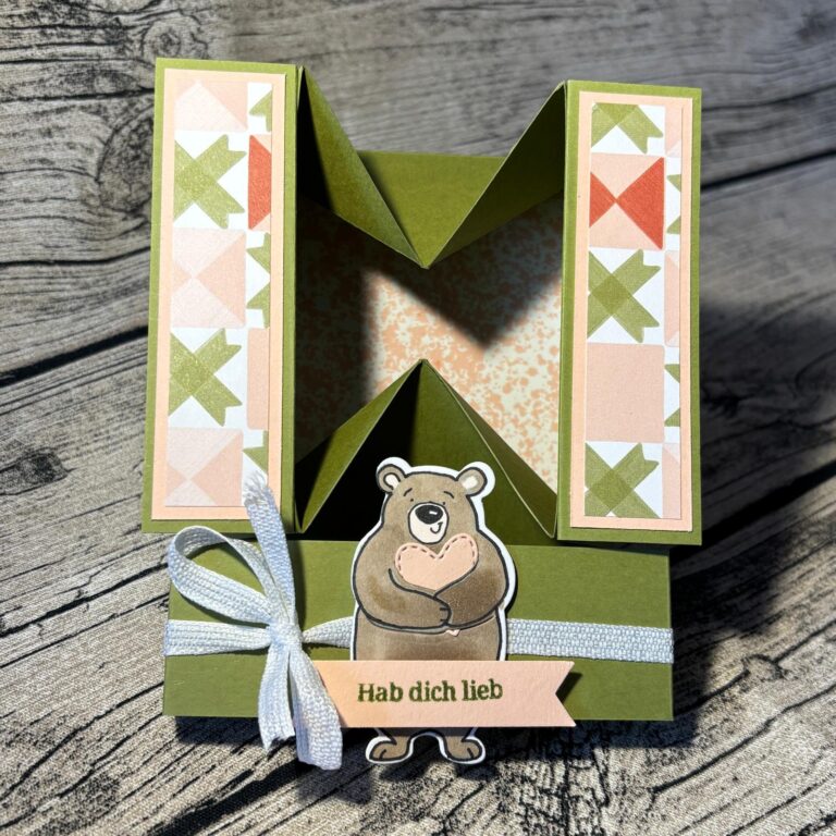 Eine handgefertigte grüne Karte, inspiriert von Mach was draus 26. Februar, zeigt Quiltmuster, einen Bären, der ein Herz hält, ein weißes Band und ein Banner mit der Aufschrift "Hab dich lieb".