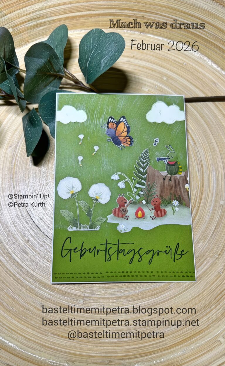 Eine handgefertigte Geburtstagskarte mit Waldtieren, Blumen und einem Schmetterling, fotografiert auf einer Holzfläche mit grünen Blättern daneben. Der Text lautet: "Mach was draus Februar 26" und Grüße auf Deutsch.
