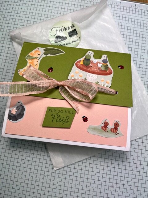 Eine handgefertigte Karte mit Tier- und Pilzaufklebern, einer rosafarbenen Schleife und dem Text FÜR SO VIEL FLEIß" auf Millimeterpapier neben einer Seidenpapierhülle mit der Aufschrift Februar" für Mach war draus 26. Februar.