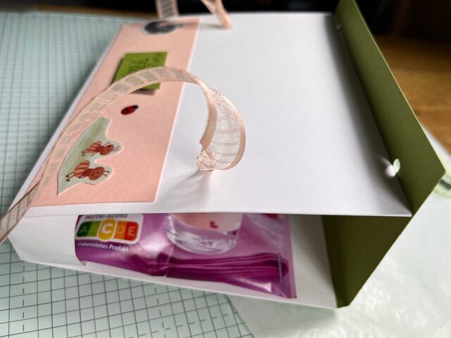 Eine Geschenktüte aus handgeschöpftem Papier mit Henkeln aus rosa Schleifenband und dekorativen Aufklebern mit dem Motiv "Mach was draus Februar 26". Ein sichtbares verpacktes Produkt lugt hervor, während die Tüte auf einer gitterartigen Oberfläche ruht.