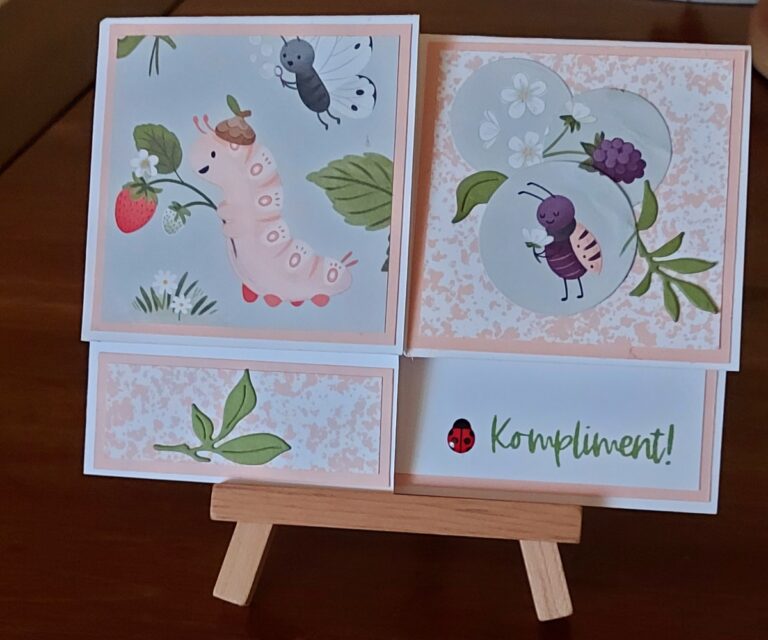 Eine handgefertigte Karte auf einer kleinen hölzernen Staffelei zeigt Cartoon-Insekten, Früchte und Blätter, mit dem Wort "Kompliment!" und einem Marienkäfer auf dem unteren rechten Feld - perfekt für Mach was draus 26. Februar.