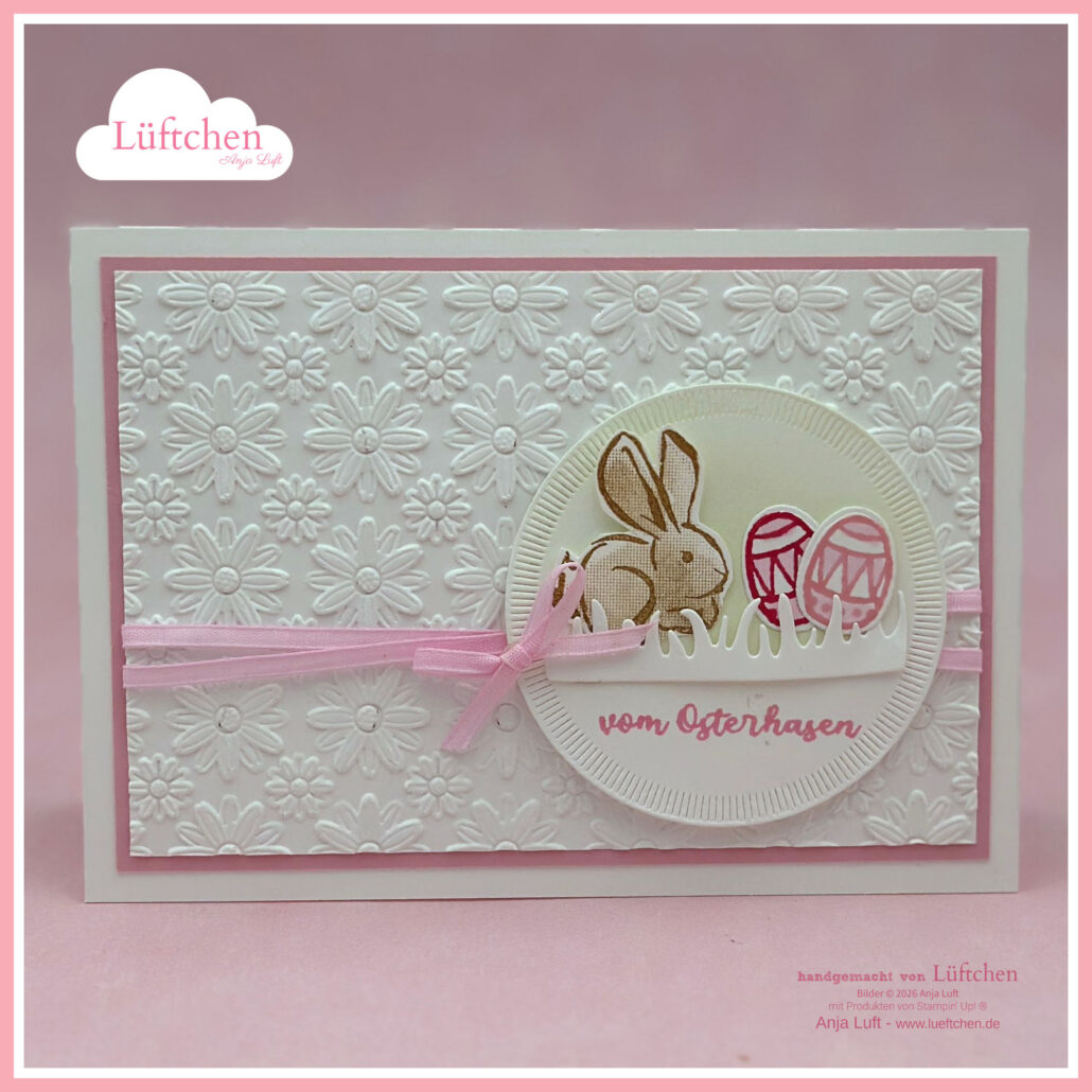 Eine handgefertigte Osterkarte für Ostern mit geprägten weißen Blumen, einer rosa Schleife und einer runden Illustration eines Hasen mit verzierten Eiern sowie dem Text "vom Osterhasen". Perfekt, um den Charme des Frühlings zu feiern.