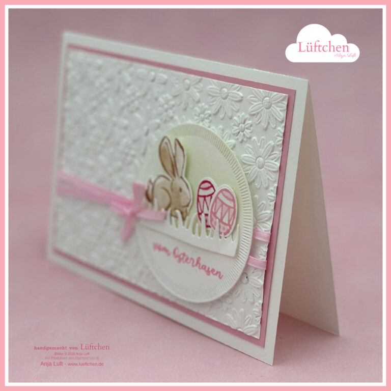 Eine handgefertigte Osterkarte mit geprägten weißen Blumen, einem rosafarbenen Band und einem runden Ausschnitt mit einem Hasen und Ostereiern vor einem rosafarbenen Hintergrund - perfekt für die Feier von Frühling/Ostern.