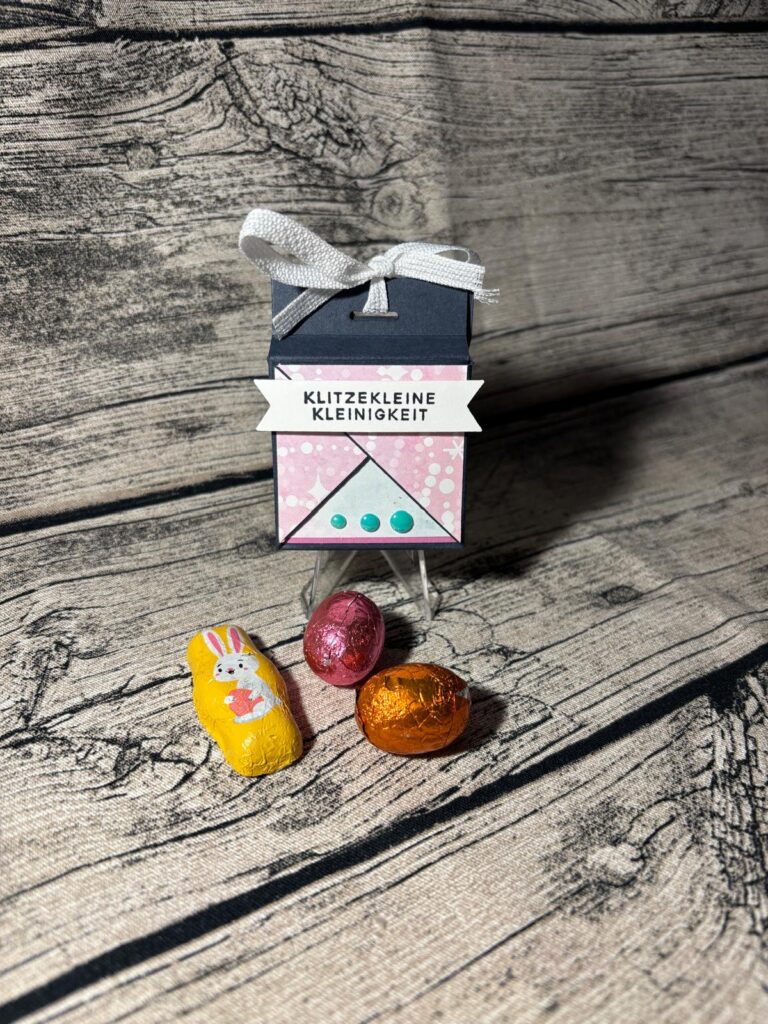 Eine kleine Geschenkschachtel mit einer Schleife und dem deutschen Text "Mach was draus März 26" steht neben drei in Folie verpackten Schokoladenpralinen, eine davon mit einem Hasenmotiv, auf einer Holzfläche.
