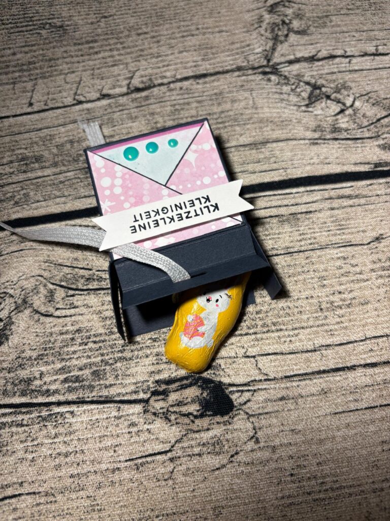 Eine kleine verzierte Erdnuss, die als Katze bemalt ist, liegt teilweise in einer schwarzen Papierschachtel mit einem Band und einem deutschen Etikett mit der Aufschrift "Kleiner Zeitvertreib" - eine charmante Mach was draus März 26 Kreation.