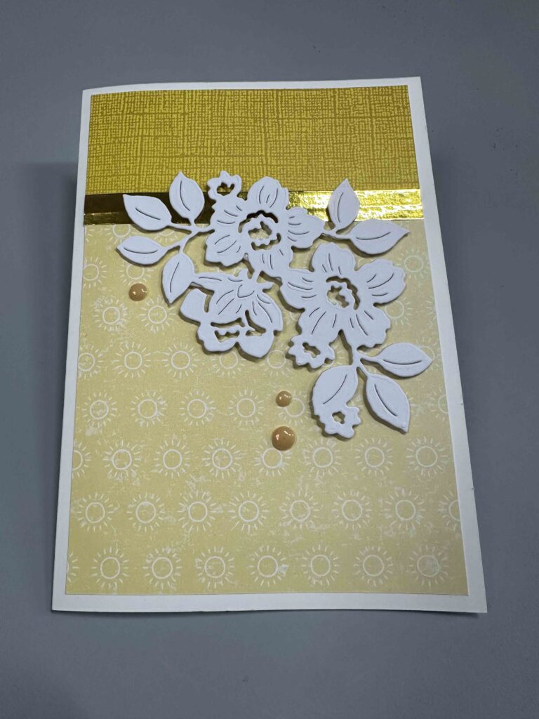 Eine dekorative Grußkarte von Mach war draus März 26, mit einem gelb gemusterten Hintergrund, einem goldenen Akzentstreifen und eleganten weißen Papierblumenausschnitten auf der Vorderseite.