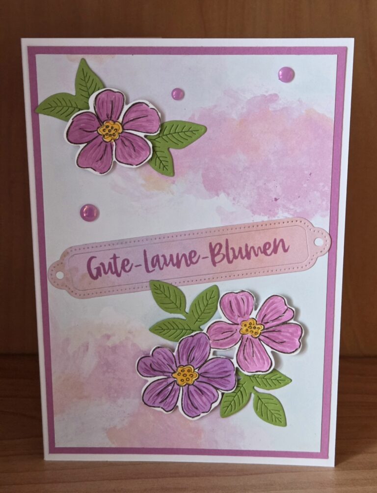 Eine Grußkarte mit rosa und lila Blumen, grünen Blättern und dem deutschen Text "Gute-Laune-Blumen" auf einem rosa und weißen Aquarellhintergrund - perfekt für Mach was draus 26. März.