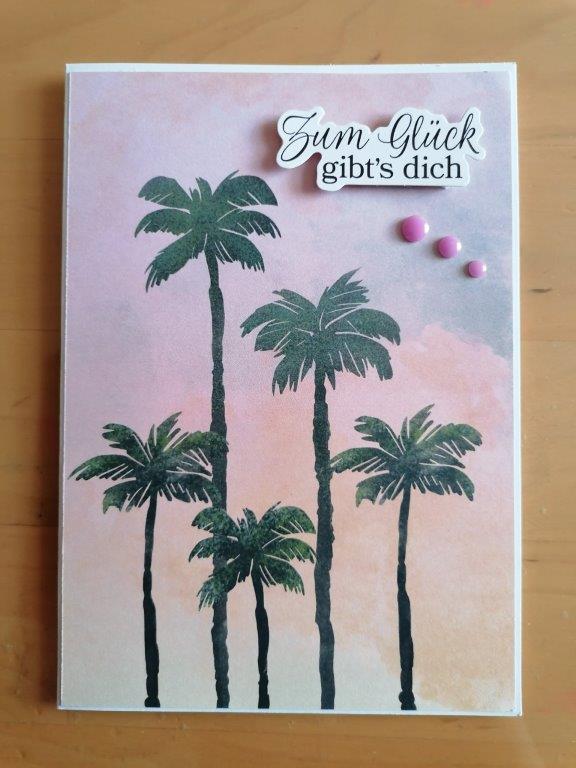 Eine Grußkarte von Mach was draus 26. März mit illustrierten Palmen vor einem rosa und lavendelfarbenen Himmel, mit dem deutschen Text "Zum Glück gibt's dich" und drei rosa Zierpunkten.