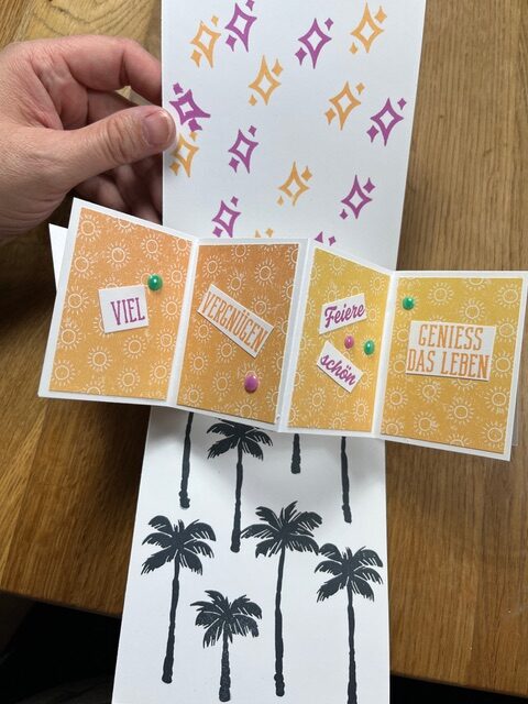 Eine Hand hält eine aufgeklappte Grußkarte, die mit orangefarbenen Mustern, dem deutschen Text "Mach was draus März 26", bunten Stecknadeln und schwarzen Palmenabbildungen auf einem Holztisch verziert ist.