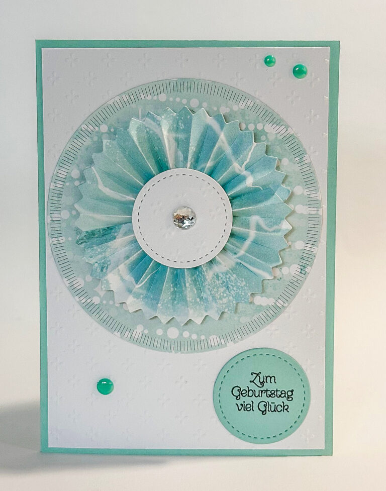 Handgefertigte Grußkarte, inspiriert von Mach was draus 26. März, mit einer blau-weißen, kreisförmig gefalteten Papierrosette, einem Edelstein in der Mitte und dem deutschen Text "Zum Geburtstag viel Glück" auf einem grün umrandeten, weißen Hintergrund.