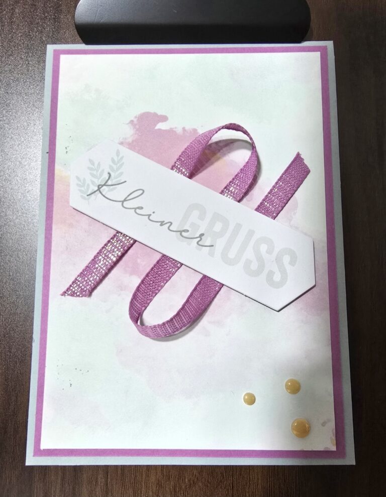 Eine handgefertigte Grußkarte mit einer rosa Schleife, Aquarell-Hintergrund und einem "Kleiner Gruss"-Anhänger auf einer Holzoberfläche - perfekt für Mach was draus 26. März.