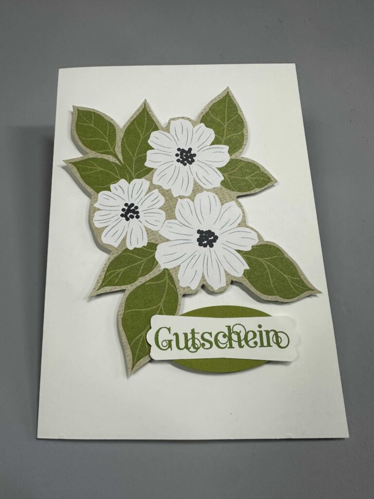 Eine weiße Karte, die mit grünen Blättern und weißen Blumen verziert ist und auf der unten das Wort "Gutschein" aufgedruckt ist - perfekt für Mach war draus 26. April.