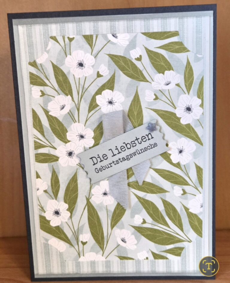 Eine blumengemusterte Geburtstagskarte in Deutsch sagt "Die liebsten Geburtstagswünsche" mit einem grauen Band und weißen Blumen auf einem grünen und blauen Hintergrund - perfekt für Mach war draus 26. April.