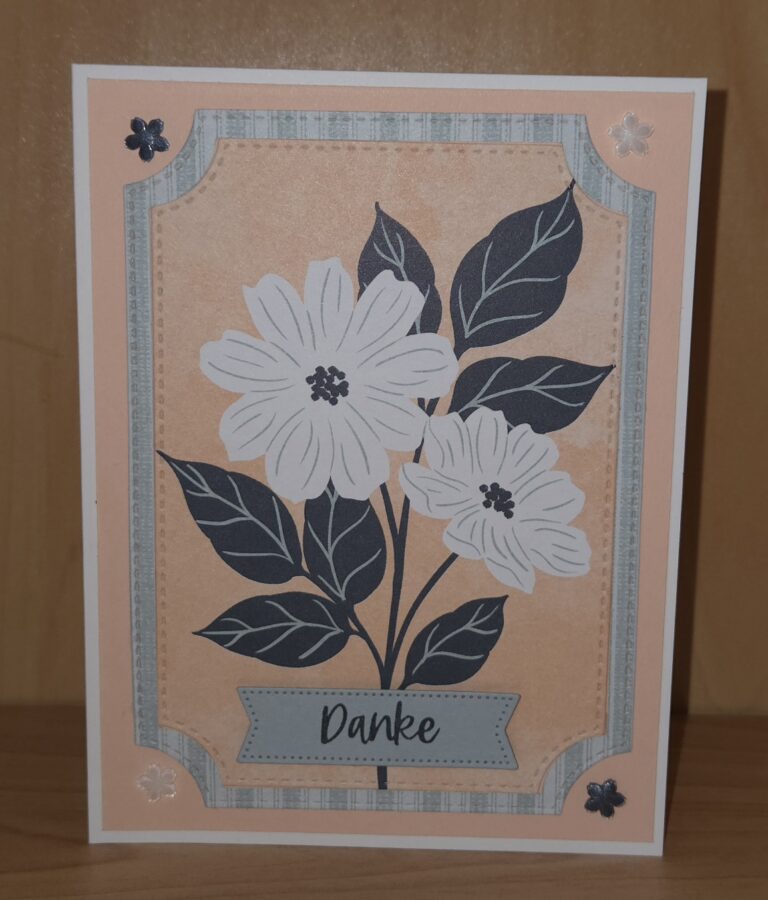 Eine Grußkarte mit weißen und dunkelblauen Blumen, grünen Blättern und "Danke" auf einem Banner, vor einem pfirsichfarbenen Hintergrund mit dekorativen Ecken - perfekt für Mach was draus 26. April Inspiration.