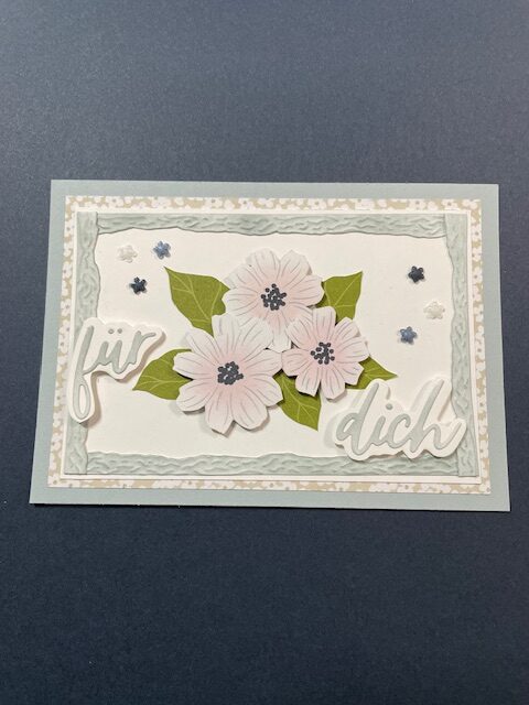 Eine handgefertigte Karte, die von Mach inspiriert ist, wurde am 26. April draus gemacht. Sie zeigt drei weiße Blumen, grüne Blätter, dekorative Umrandungen und die deutschen Worte "für dich" auf einem blau-grauen Hintergrund.