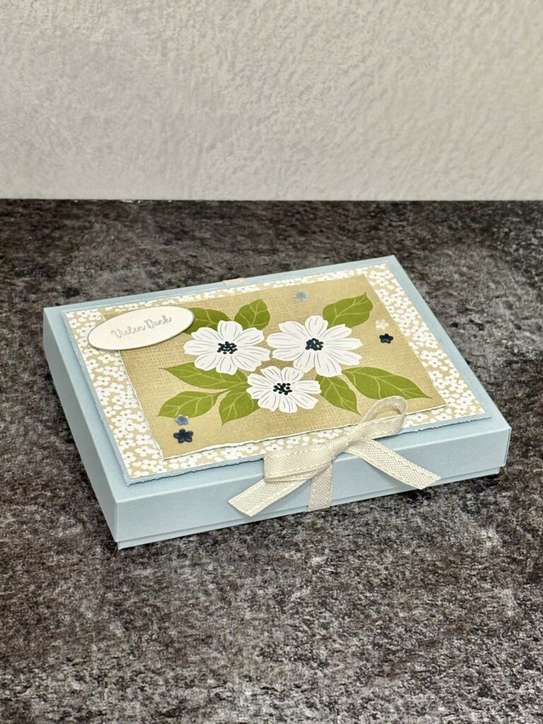 Ein hellblauer, rechteckiger Geschenkkarton mit cremefarbenem Band, verziert mit einem Blumenmuster und einem kleinen ovalen Anhänger mit der Aufschrift "Mit besten Grüßen", perfekt zum Feiern von Mach wurde am 26. April draus.