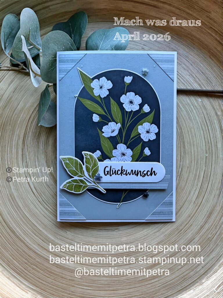 Eine handgefertigte Grußkarte mit weißen Blumen und grünen Blättern auf dunklem Hintergrund, beschriftet mit "Glückwunsch". Der Text hebt einen Blog und das Projekt "Mach was draus 26. April" hervor.