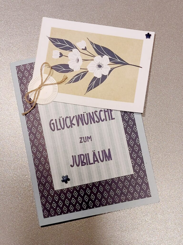 Eine Grußkarte mit der Aufschrift "Glückwünsche zum Jubiläum" auf Deutsch, gekrönt von einer kleinen, mit Schnur gebundenen Blumenkarte, inspiriert von Mach was draus 26. April, auf einer strukturierten Oberfläche platziert.