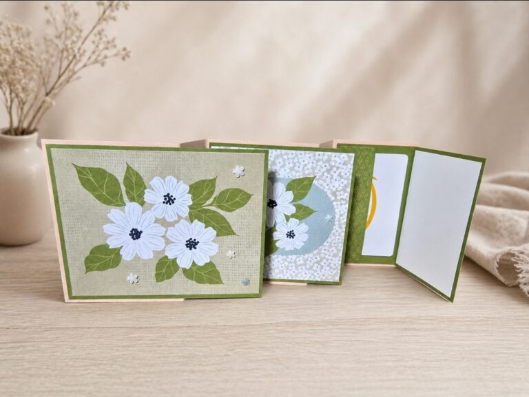 Eine handgefertigte Grußkarte im Ziehharmonika-Stil mit floralen Motiven aus weißen Blüten und grünen Blättern, die auf einer hölzernen Oberfläche präsentiert wird - perfekt für die Feier von Mach war draus 26. April.
