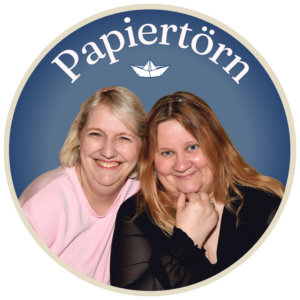 Anja und Petra sitzen lächelnd dicht beieinander vor einem blauen, runden Hintergrund, über dem das Wort „Papiertörn“ und die Abbildung eines Papierboots zu sehen sind, und feiern den bevorstehenden Papiertörn-Basteltag 2026.